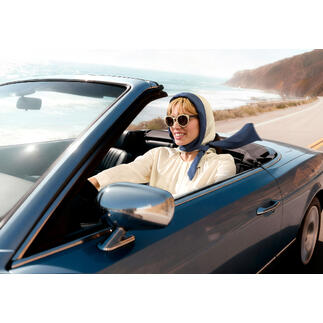 Frau in einem Cabrio mit Kopftuch und Sonnenbrille fährt an Küste entlang.