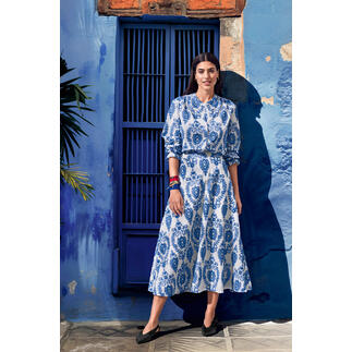 Frau in einem blauen Kleid mit weißem Muster, steht vor einer blauen Wand.