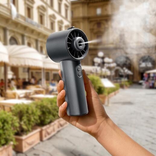 Hand hält einen Hydro-Mini-Ventilator in einer Stadt, mit historischen Gebäuden im Hintergrund.