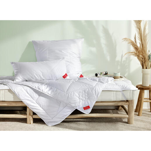 Bett mit weißen Kissen und einer Decke, helle Schlafzimmeratmosphäre.