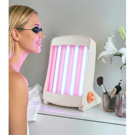 Gesichtsbräuner mit pinken und blauen Lampen, steht auf einem Tisch neben Kosmetikartikeln.