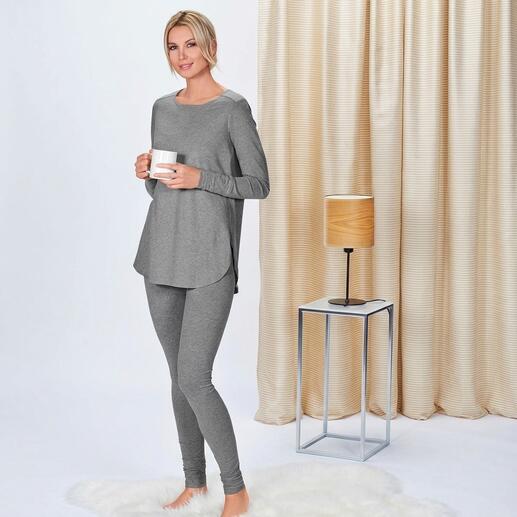 Frau in grauem Longshirt und Sweatpants hält eine Tasse, stehend in einem modernen Raum.
