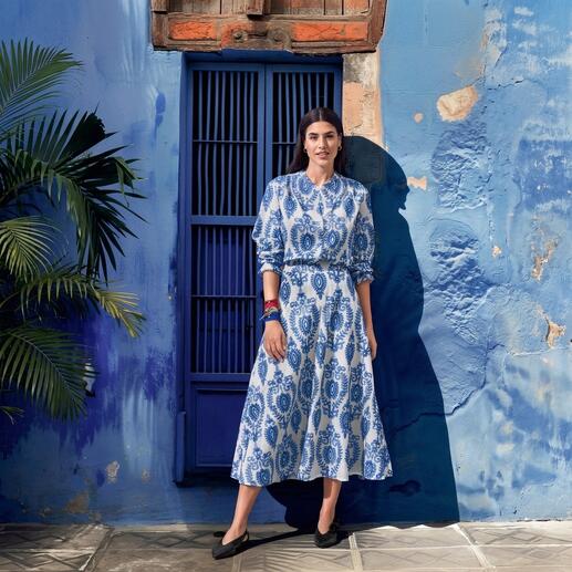 Frau in einem blauen Kleid mit weißem Muster, steht vor einer blauen Wand.