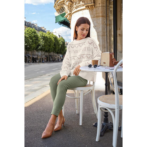 Pourchet Paris Raffia-Boxy-Bag Frau in gestricktem Pullover sitzt an einem Tisch mit einem Kaffee und einer Tasche, mit Stadt im Hintergrund.
