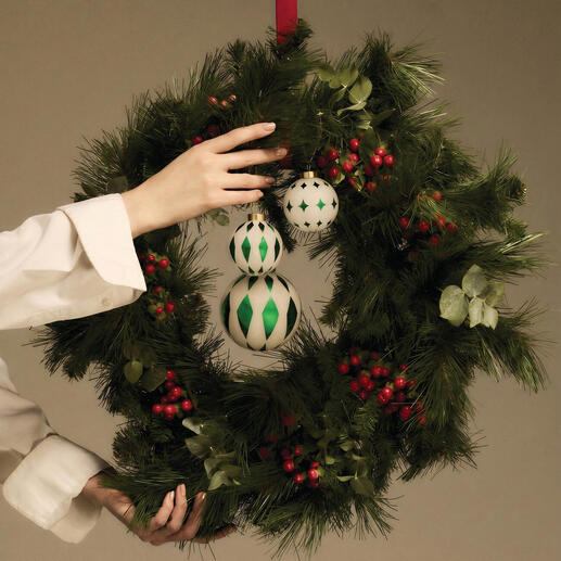 Hand mit Weihnachtskugeln über einem grünen Kranz mit roten Beeren.