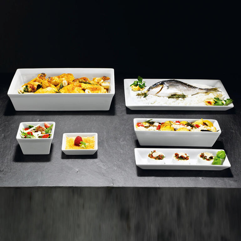 Verschiedene weiße Geschirrformen auf einem dunklen Tisch, mit unterschiedlichem Essen.