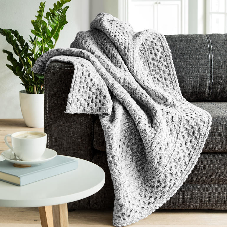 Aran-Strickplaid in hellem Grau, auf einem Sofa drapiert, mit einer Tasse Kaffee.