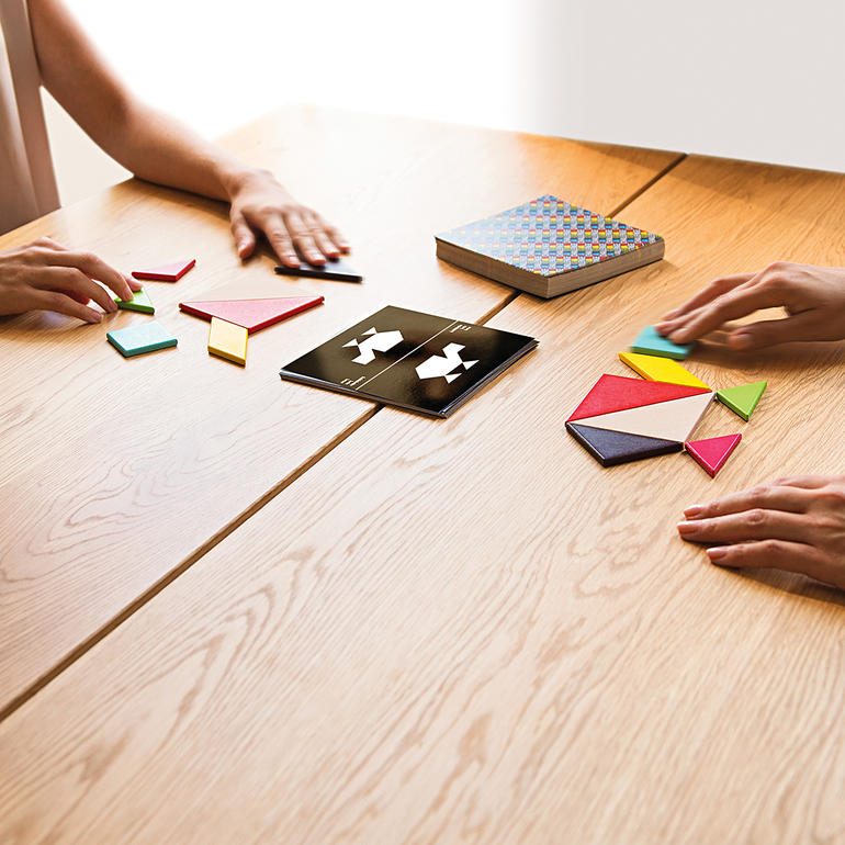 Zwei Personen spielen Tangram auf einem Tisch mit Holzoberfläche.