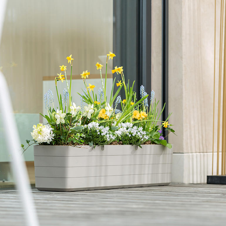 Heller Blumenkasten mit verschiedenen Blumen, platziert auf einer Holzterrasse.