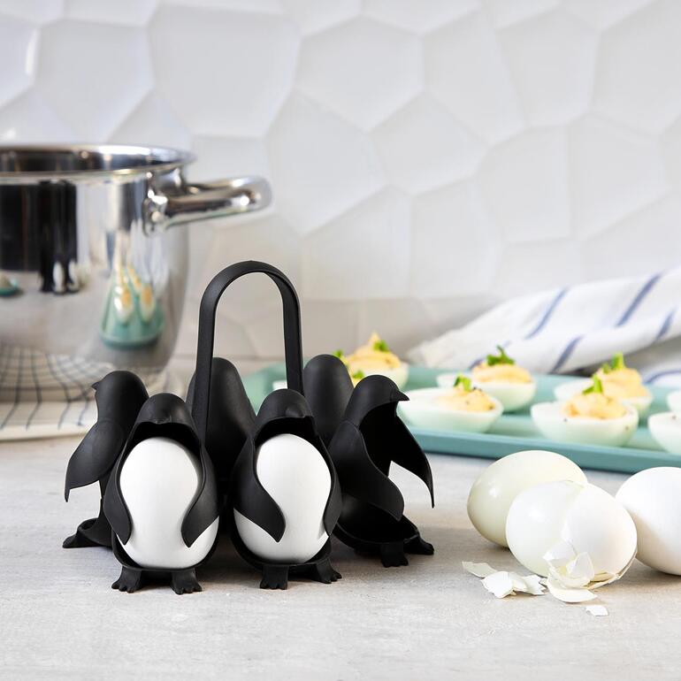 Eierhalter in Form von Pinguinen mit gekochten Eiern und einer Topfdekoration im Hintergrund.