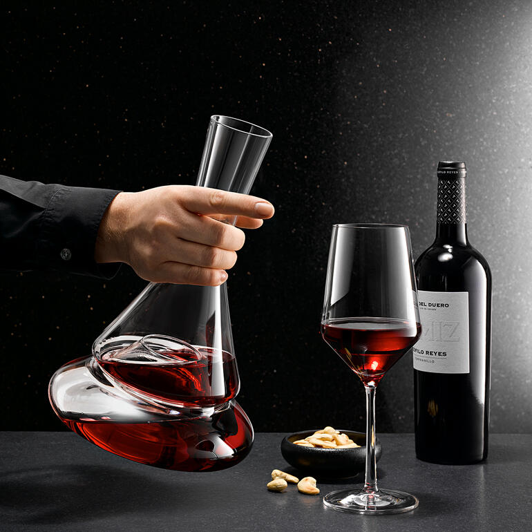 Doppio Doppelstock-Dekanter mit Rotwein in der Hand, neben einem Rotweinglas und Nüssen.