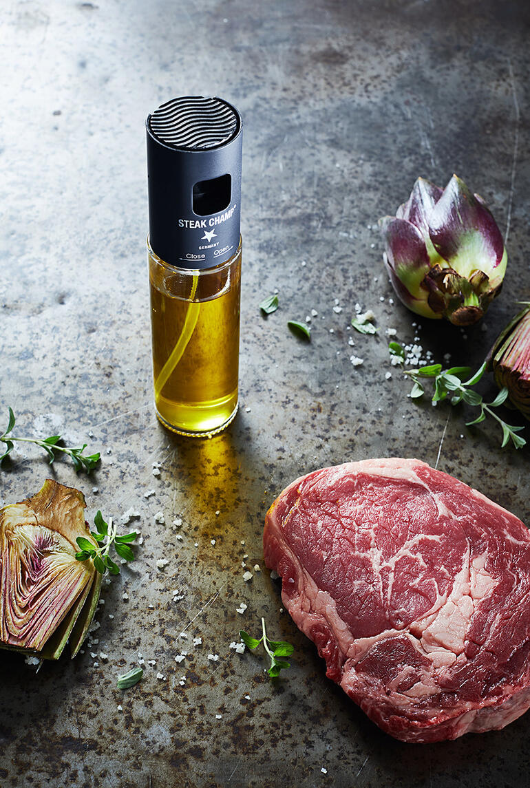 Öl-Zerstäuber mit Filter neben rohem Steak und Kunstblumen auf rustikalem Tisch.