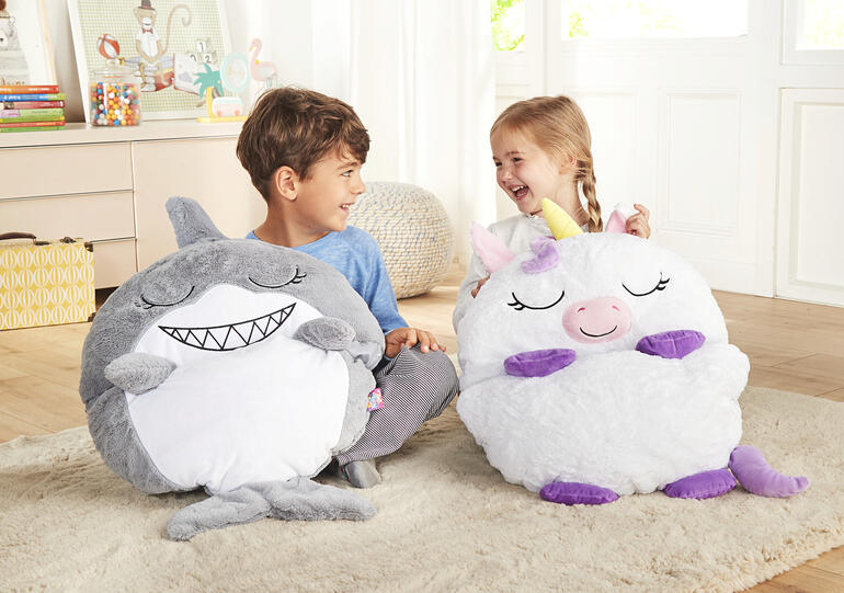 Zwei Kinder sitzen neben einem bunten Einhorn- und Hai-Kuschelschlafsack.