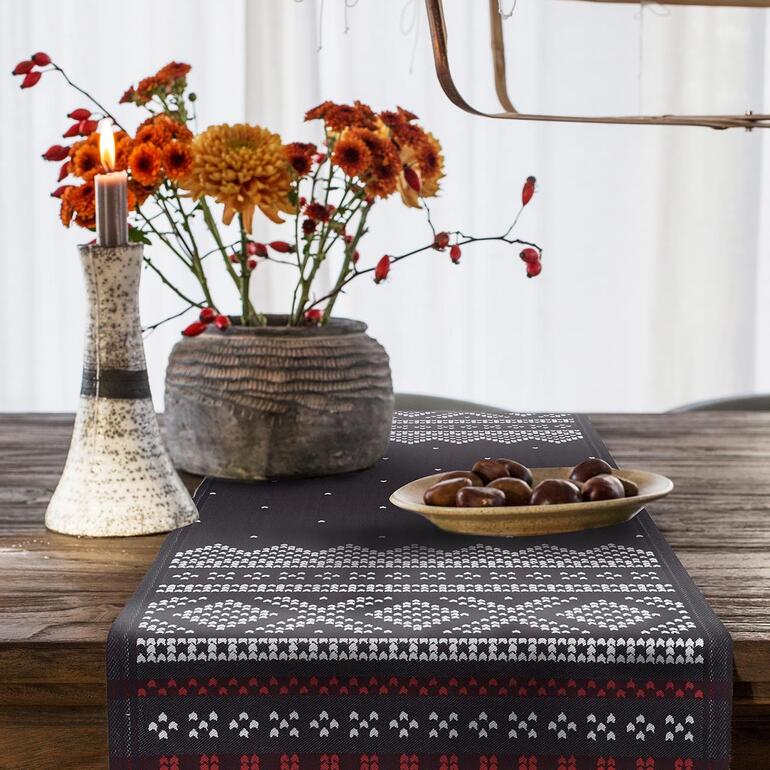 Mit sorgfältigem Einfass und materialreich schwer gewebt – so liegt der Läufer stets tadellos glatt an. Tisch mit schwarzem Tischläufer Norwegen, Kerze und Blumenstrauß.