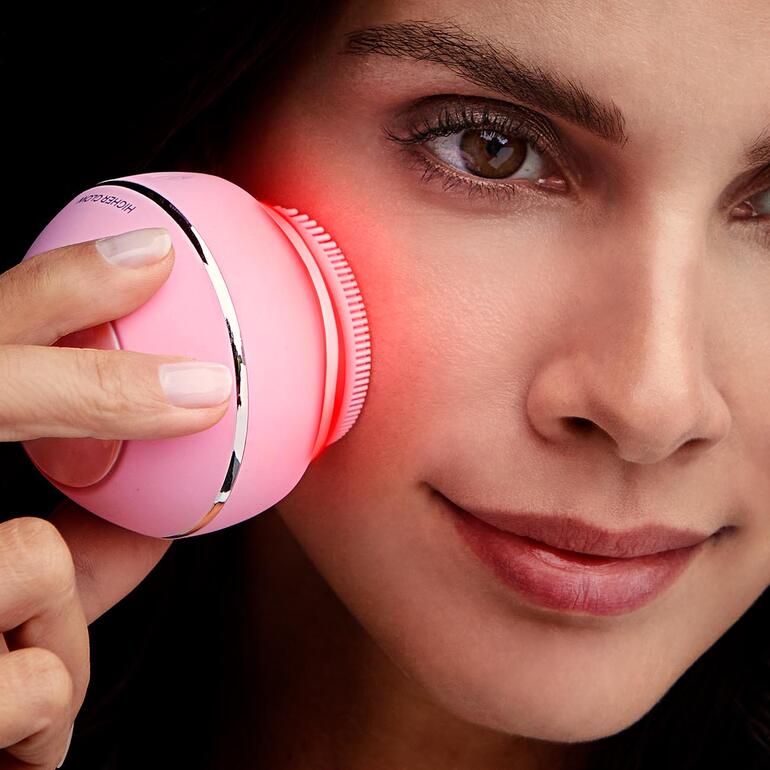 Frau mit rosa Gesichtsbürste Higher Glow in der Hand, die auf ihre Wange zielt.