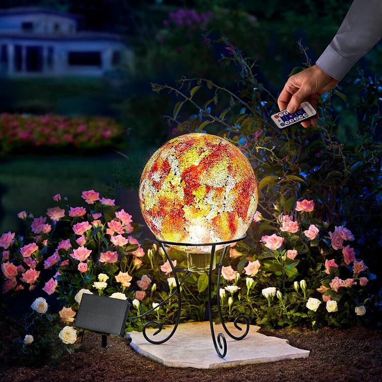 Solar-Gartenkugel auf einem Ständer, umgeben von Blumen, Hand hält Fernbedienung.