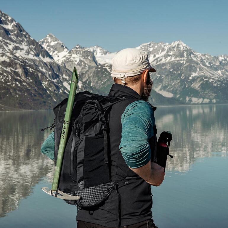 Mann mit Outdoor-Falt-Rucksack steht am Ufer eines Sees mit Bergen im Hintergrund.