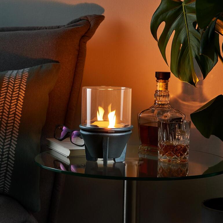 Schmelzfeuer mit Glaszylinder auf einem Tisch neben Whiskey und Pflanzen.