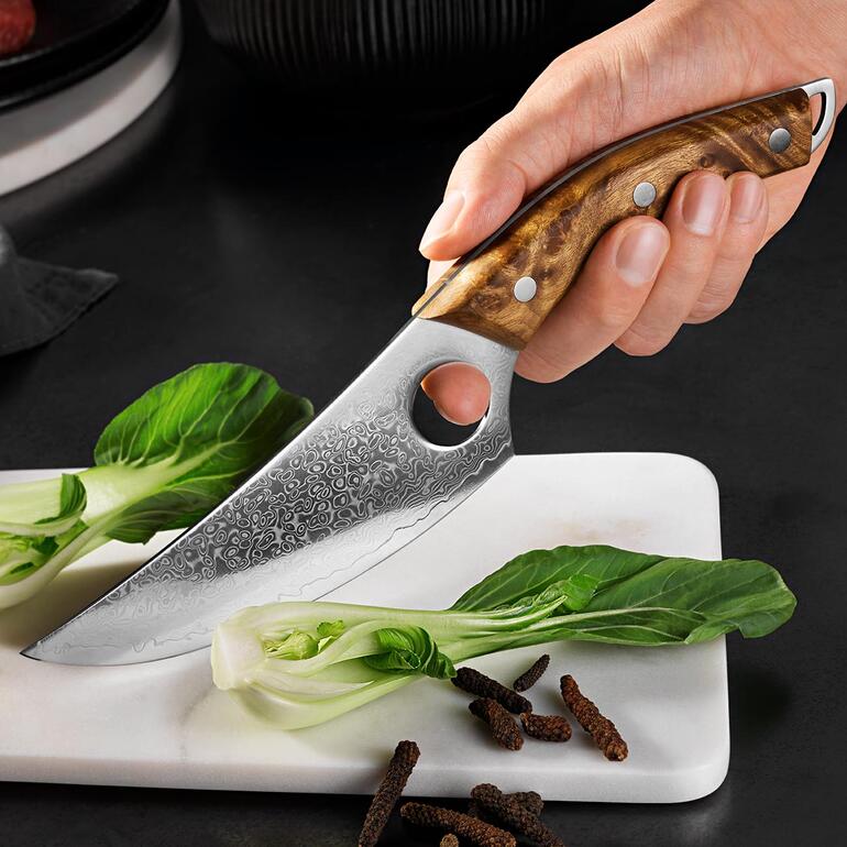 Avec trou de préhension au bout de la lame pour une prise en main sécurisée et un guidage précis. Damast Butcher-Messer in der Hand mit frischen Zutaten im Hintergrund.