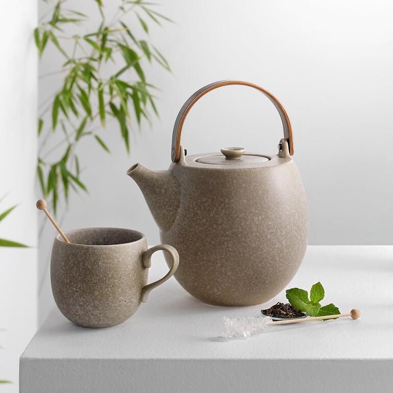 Teekanne und Becher in grauer Keramik auf einem Tisch mit einer Pflanze im Hintergrund