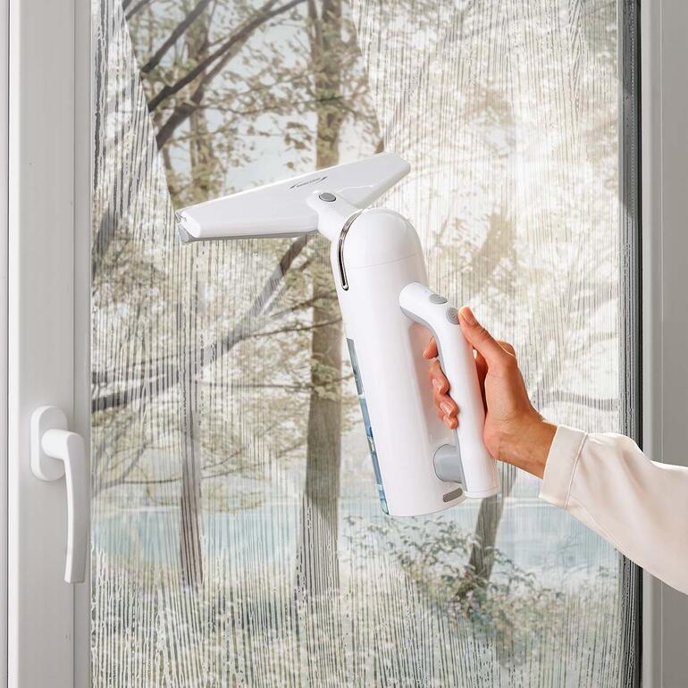 Hand hält einen weißen Akku-Fensterreiniger vor einem Fenster.