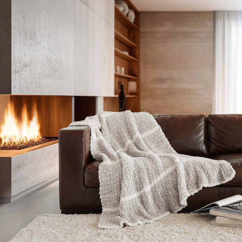 Kunstfell-Strickdecke auf einem braunen Sofa vor einem Kamin.