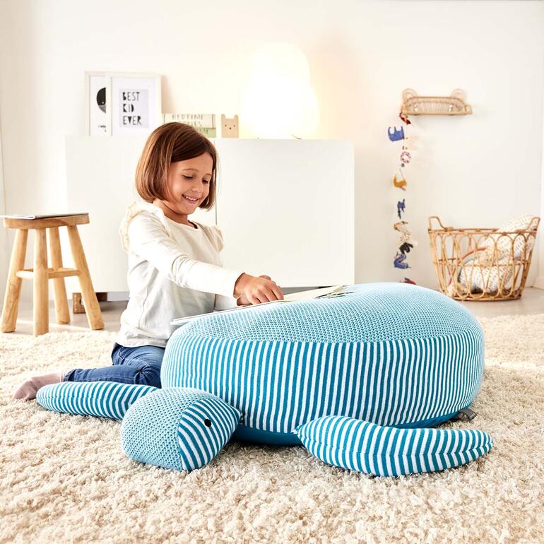 Mädchen mit Buch sitzt auf einer Kuschel-/Sitzschildkröte in einem gemütlichen Raum.