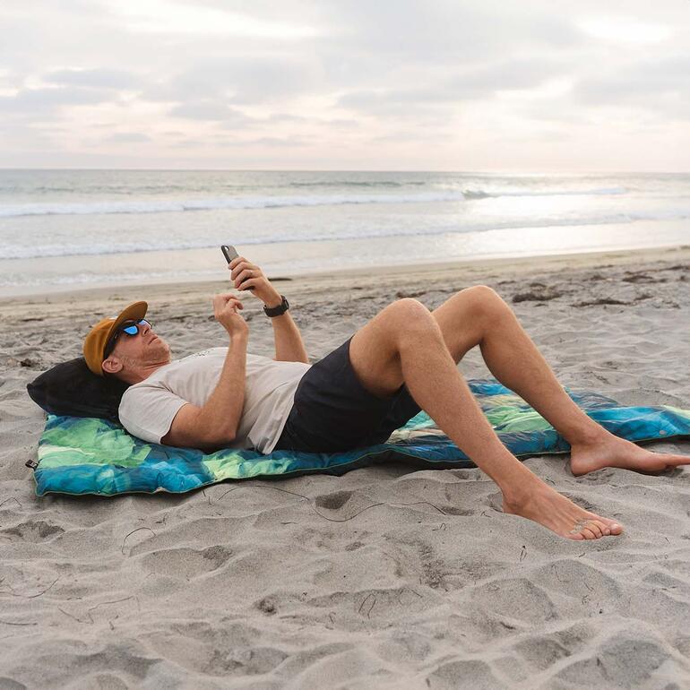 Mann liegt auf einer bunten Reisedecke am Strand und nutzt sein Smartphone