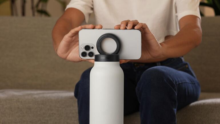 2-in-1 Trinkflasche in Schwarz mit Smartphone-Halterung, auf einem weißen Sockel.