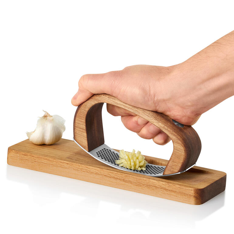 Hand hält AdHoc Knoblauchwippe über Holzbrett mit einer Knoblauchzehe.