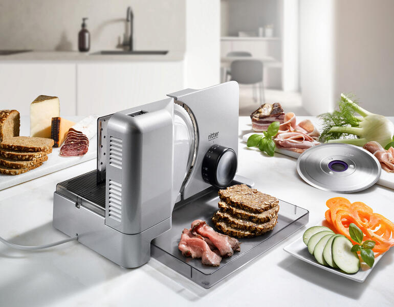 Allesschneider Duo Plus schneidet Brot und Wurst auf einer Arbeitsfläche.