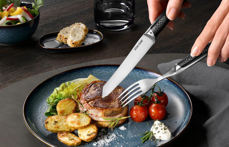 Durch die geschwungene Klingenform kommt beim Schneiden lediglich die Messerspitze mit dem Teller in Berührung – das hält die Schneide noch länger scharf. Hand, die mit Steak-Besteck Prime Rib auf einem Teller in ein Stück Steak schneidet.