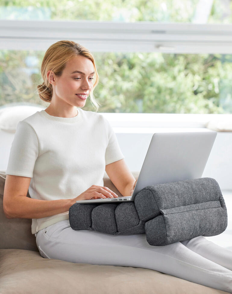 Ideal als bequemes Rückenpolster und Wirbelsäulenstütze beim Arbeiten, Autofahren, ... Frau sitzt mit Flexi-Rollenkissen, das ihre Arme unterstützt, am Laptop auf dem Sofa.