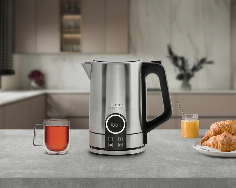 Moderner Edelstahl Wasserkocher auf einer Arbeitsplatte mit einer Tasse Tee und einem Croissant.