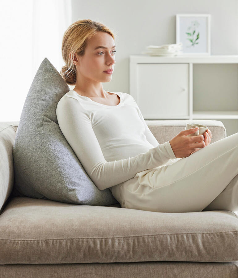 Ecru Frau sitzt mit einem ecrufarbenen Rückenkissen auf der Couch.
