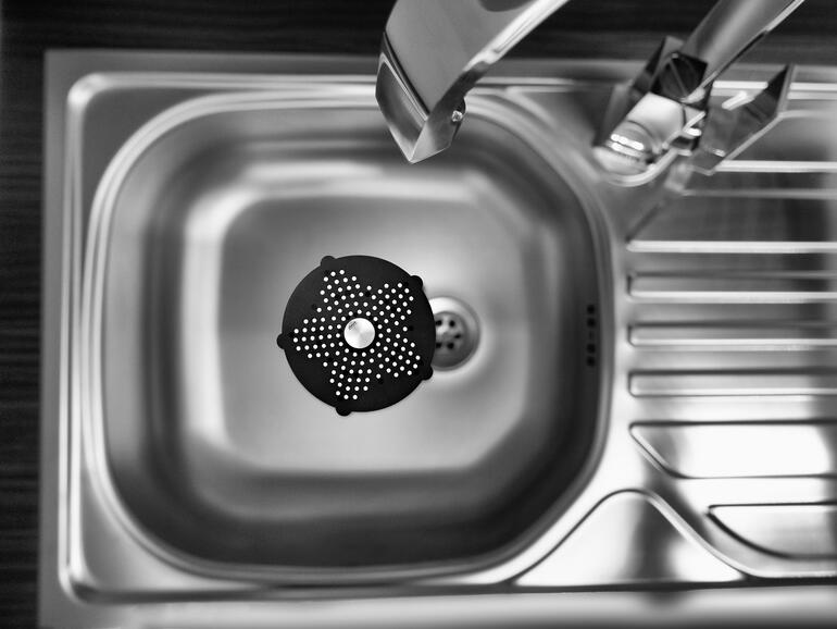 Das handtellergrosse Abflusssieb ist perfekt in der Spüle, Dusche, Wanne, über dem Bodenabfluss, ... Perspektive von oben auf ein schwarzes Geruchskiller-Abflusssieb in einer Spüle.