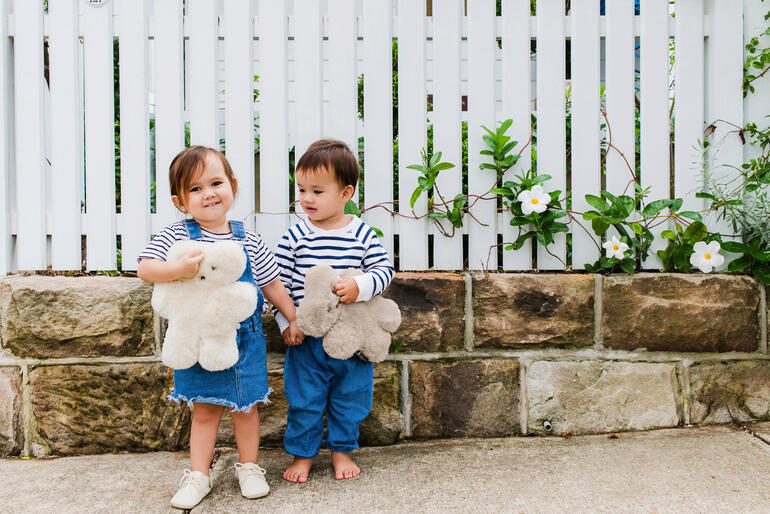 Zwei Kinder stehen an einer Mauer und lächeln, während sie Schafsfell Teddybären umarmen.