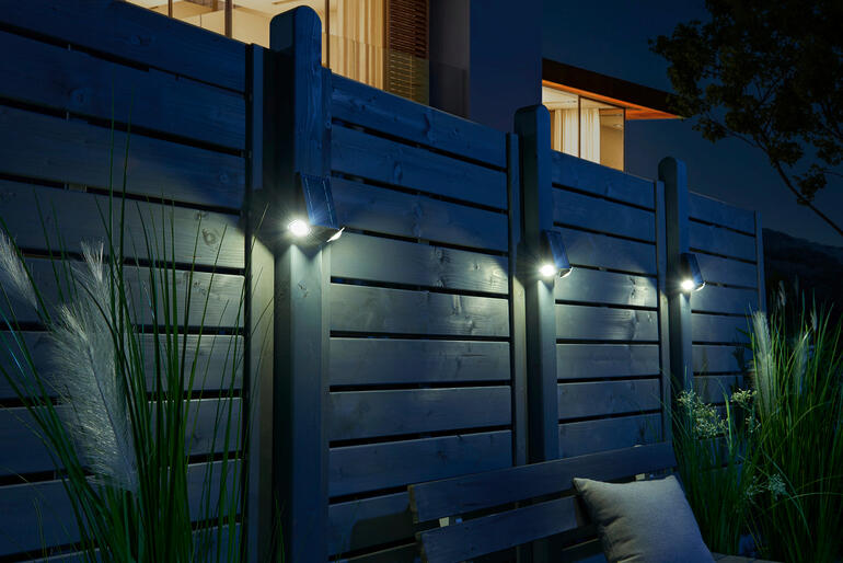 Im 3er-Set auch zum Beleuchten grösserer Bereiche geeignet. Intelligenter Solarstrahler beleuchtet eine Holzhecke bei Nacht.