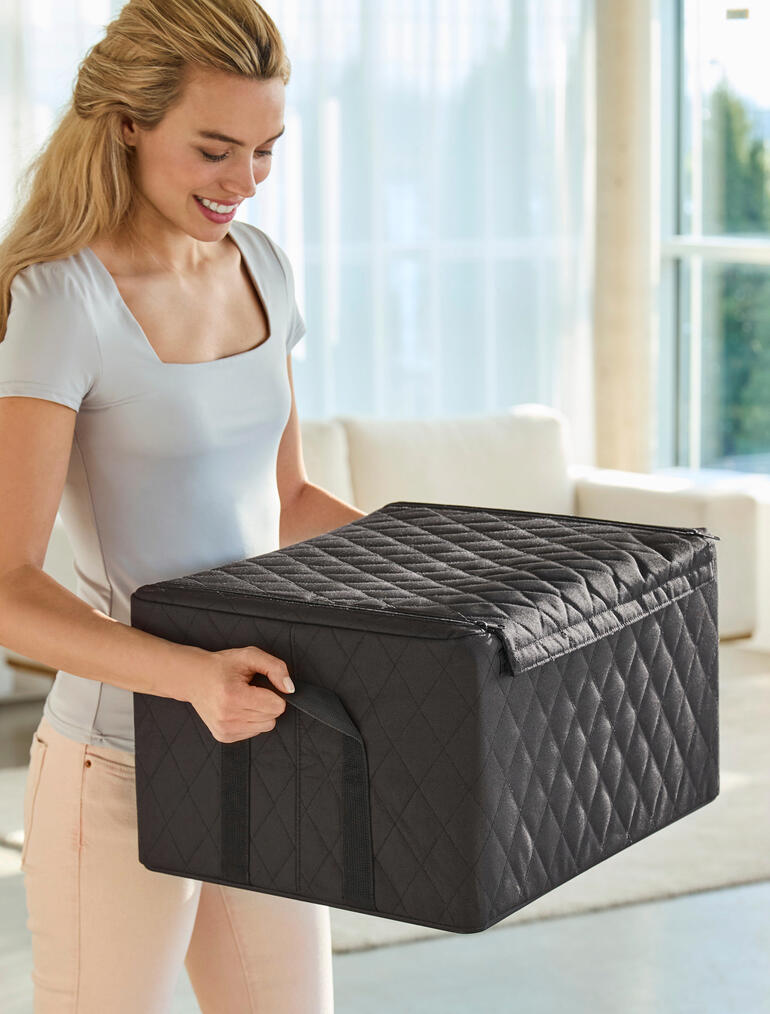 Frau hebt eine schwarze Stapelbox mit Stepp-Rautenmuster, im Wohnzimmer.