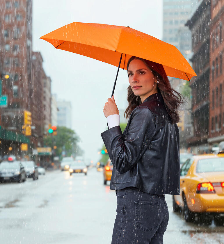 Frau hält einen orangen Taschenschirm in der Regen, auf der Straße stehend.