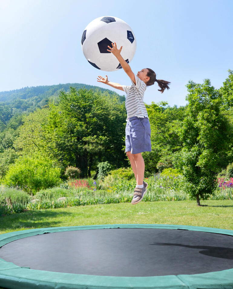 Riesiger Spielspass auch auf dem Trampolin. Mädchen springt im Garten und greift nach einem großen Fußball.