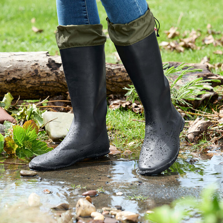 Schwarze Gummistiefel mit umgeschlagenem oberen Teil, stehen am Rand eines Wasserlaufes.