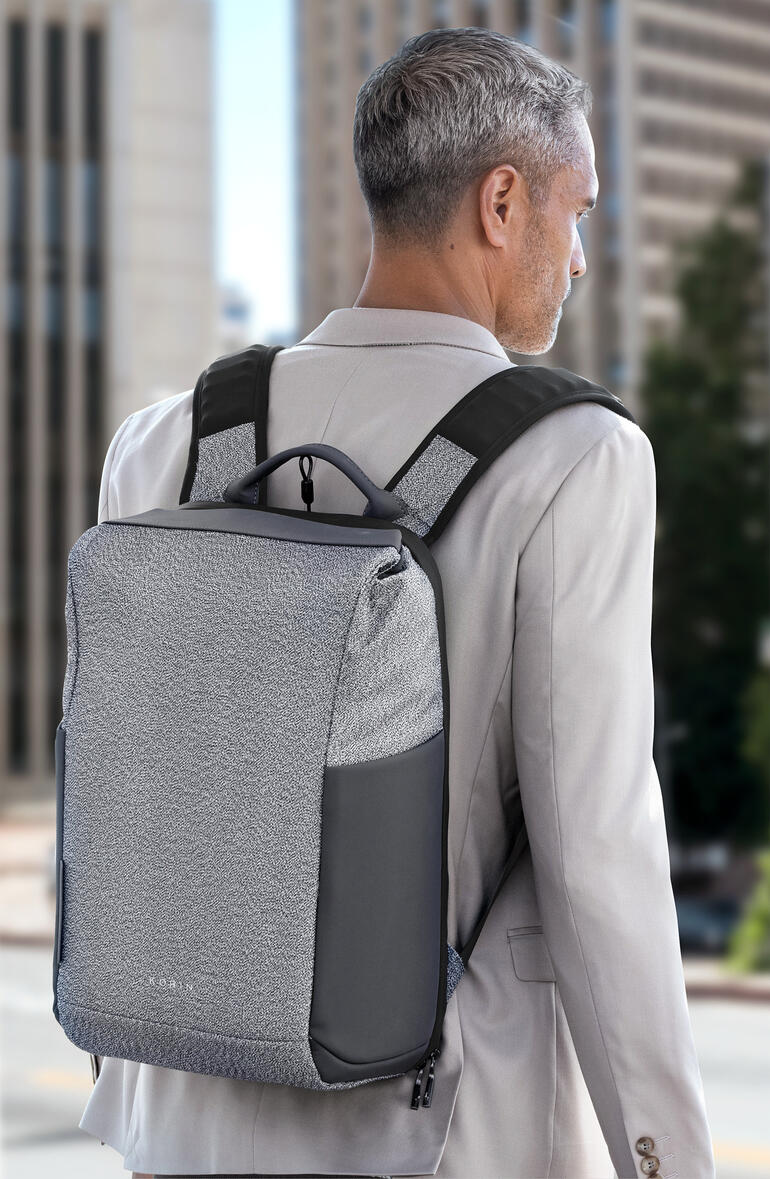 Mann mit grauem Anti-Diebstahl-Rucksack auf dem Rücken vor urbanem Hintergrund.