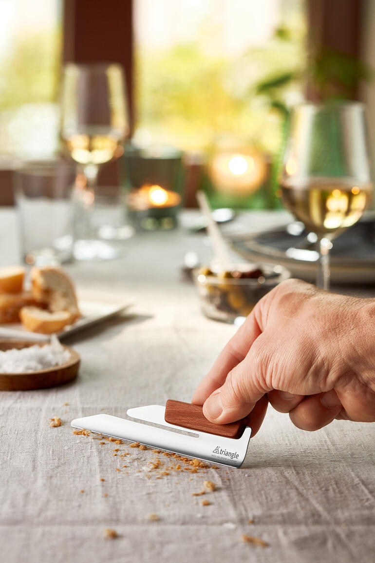 Hand zeigt den Design-Krümelsammler auf einem gedeckten Tisch mit Snacks.