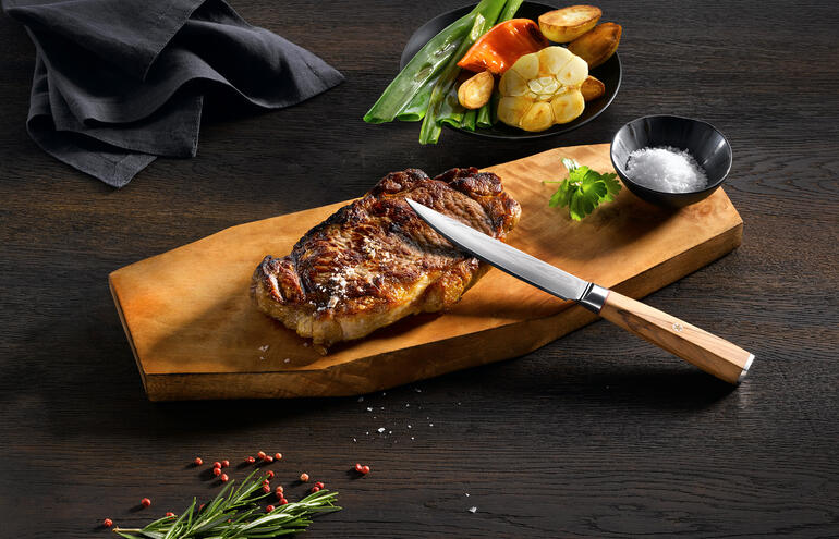Damast-Steakmesser auf Holzbrett mit gegrilltem Steak und Gemüse.
