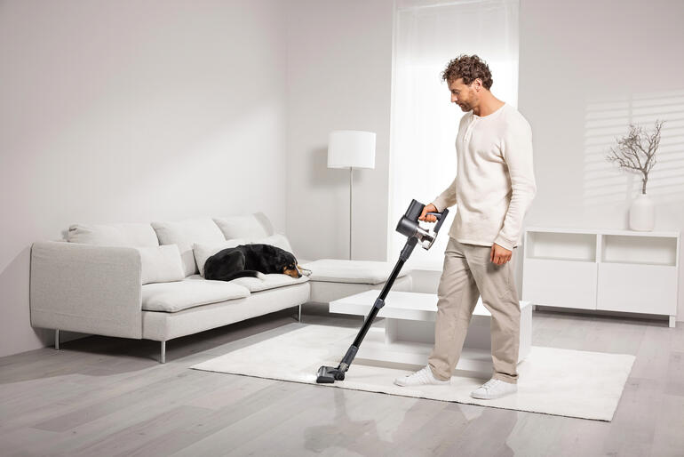 Mann saugt mit einem Akku-Staubsauger auf einem Teppich, Hund liegt auf einem Sofa.