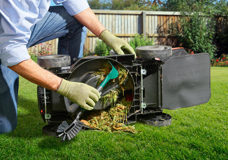 Person reinigt einen umgedrehten Rasenmäher mit einem Werkzeug, während Grasreste entfernt werden.