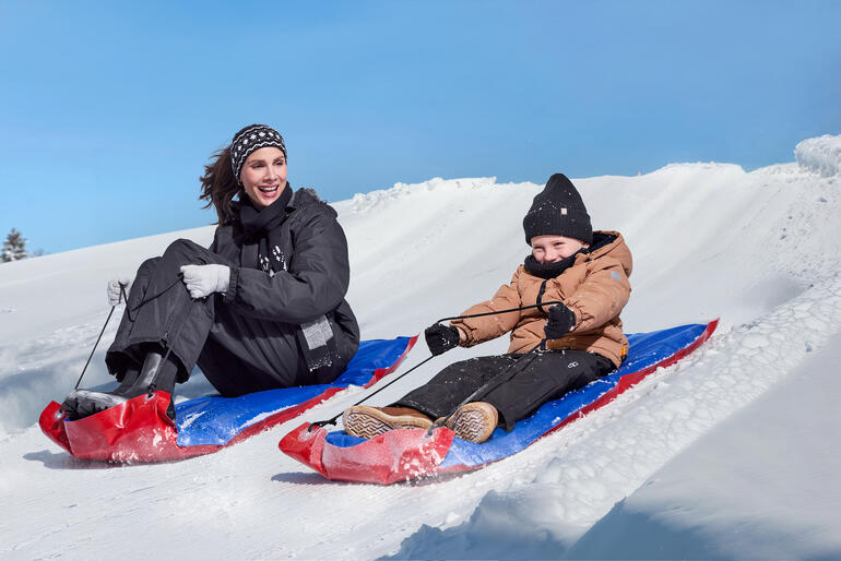 Frau und Kind fahren mit XL-Schneegleitern einen Schneehang hinunter.