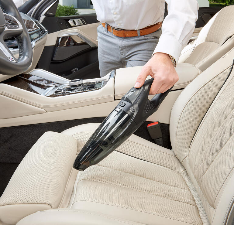 Mann benutzt einen Akku-Handsauger zur Reinigung des Autositzes mit hellem Interieur.