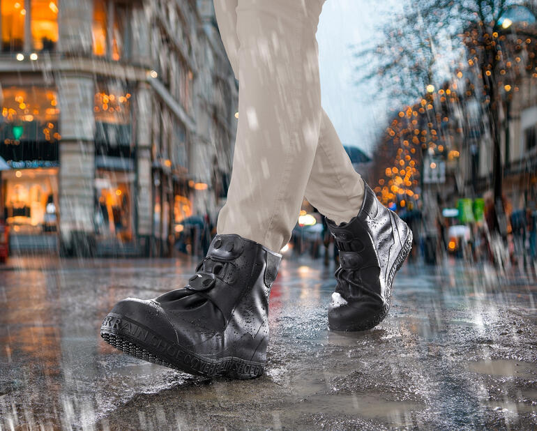 Person trägt schwarze Stiefel mit Silikon-Schuhüberziehern im Regen auf einer nassen Straße.